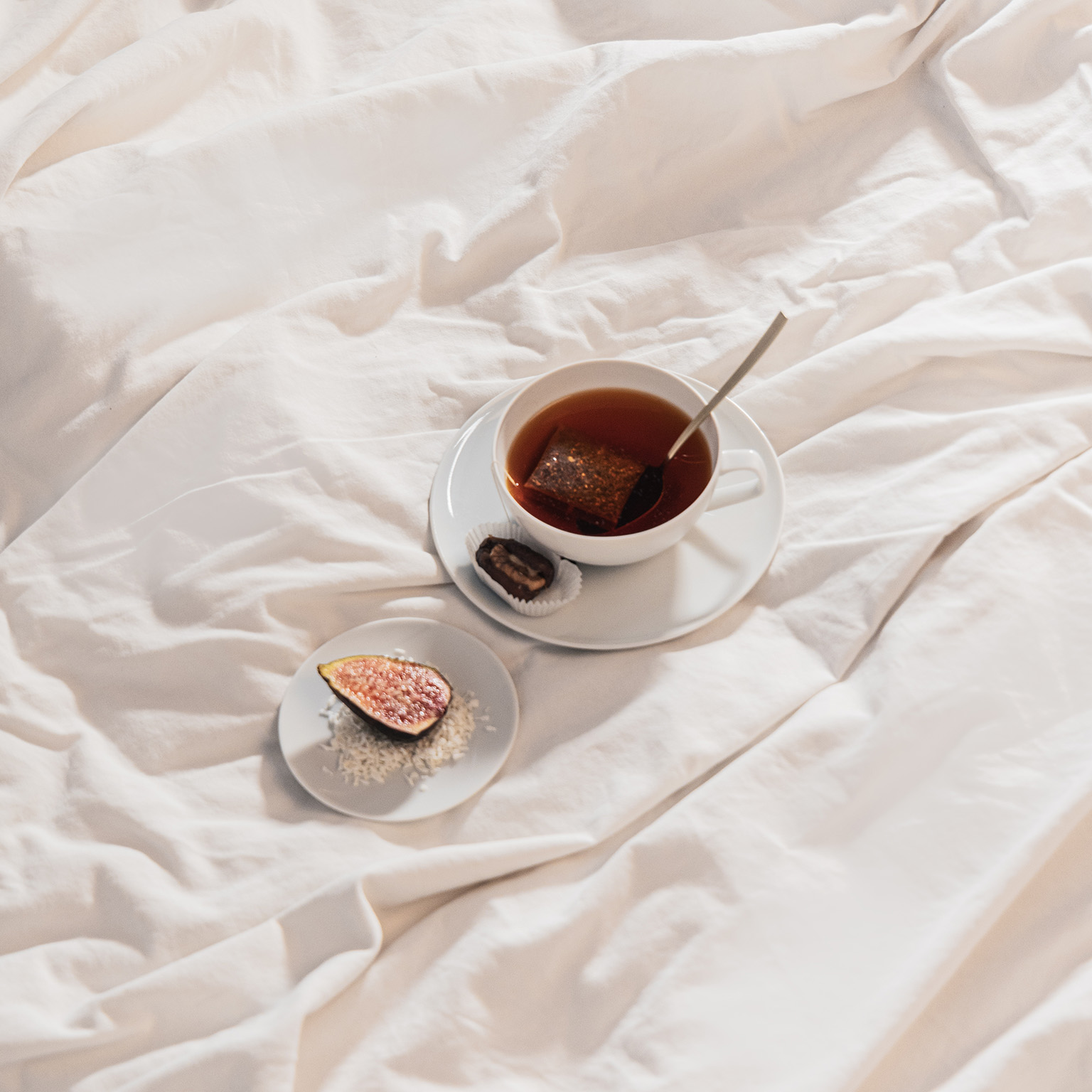 TAC tea cup filled with tea, saucer and bread plate with a fig stand on a bed sheet.